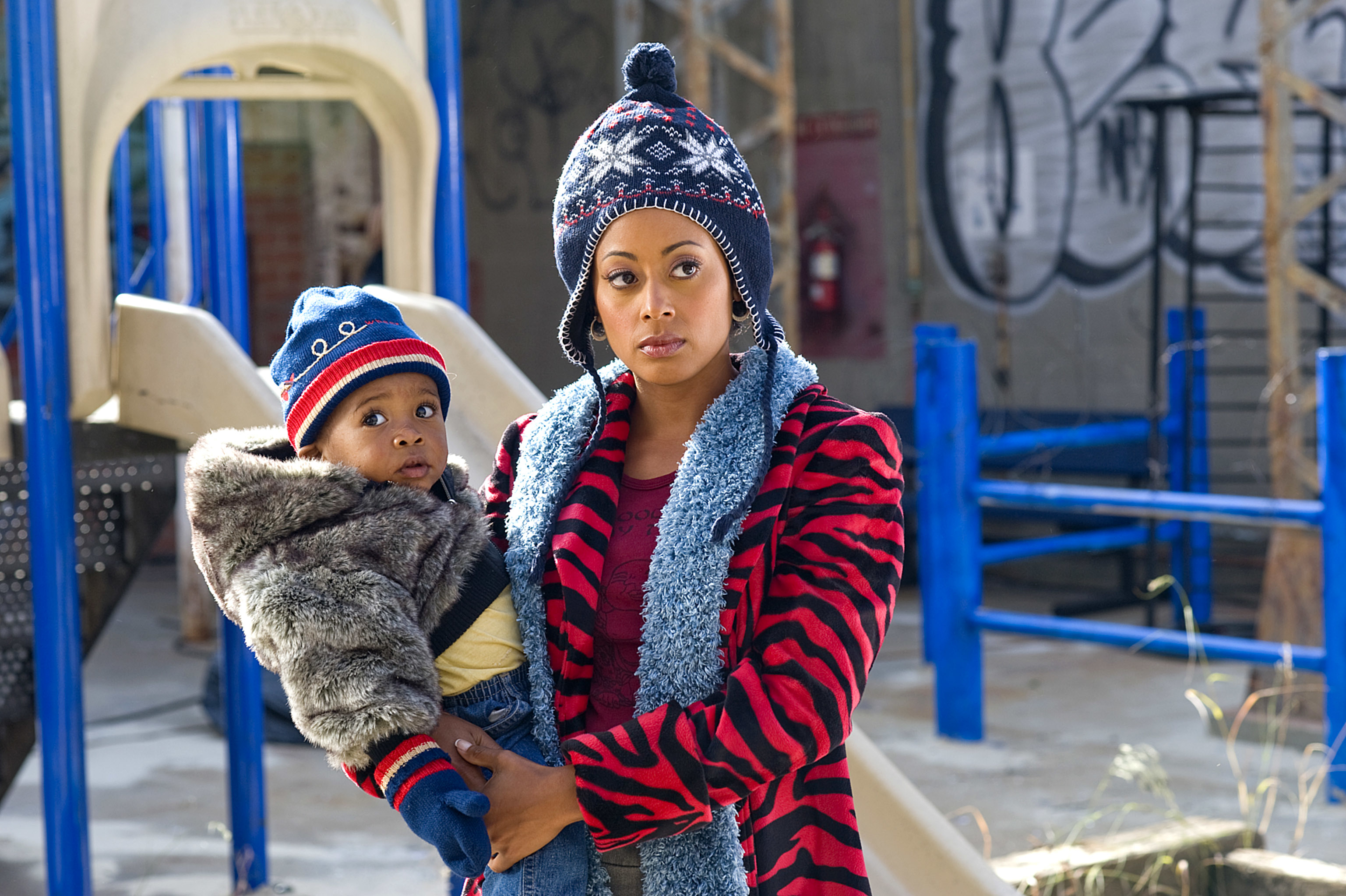 A woman in a red and black tiger-striped coat and blue scarf holds a child in her arms. Both are wearing knitted hats. They are on a playground