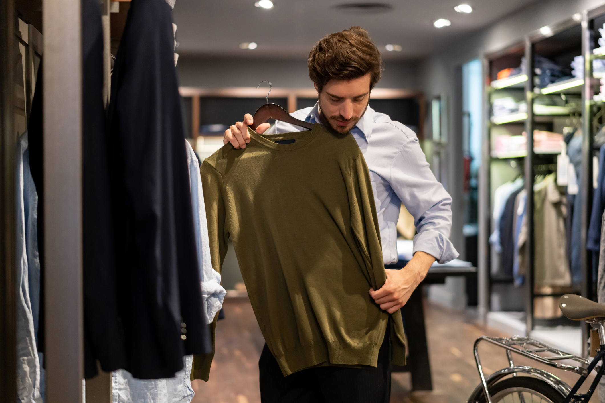 A man browses clothing in a store, holding up a sweater for closer inspection