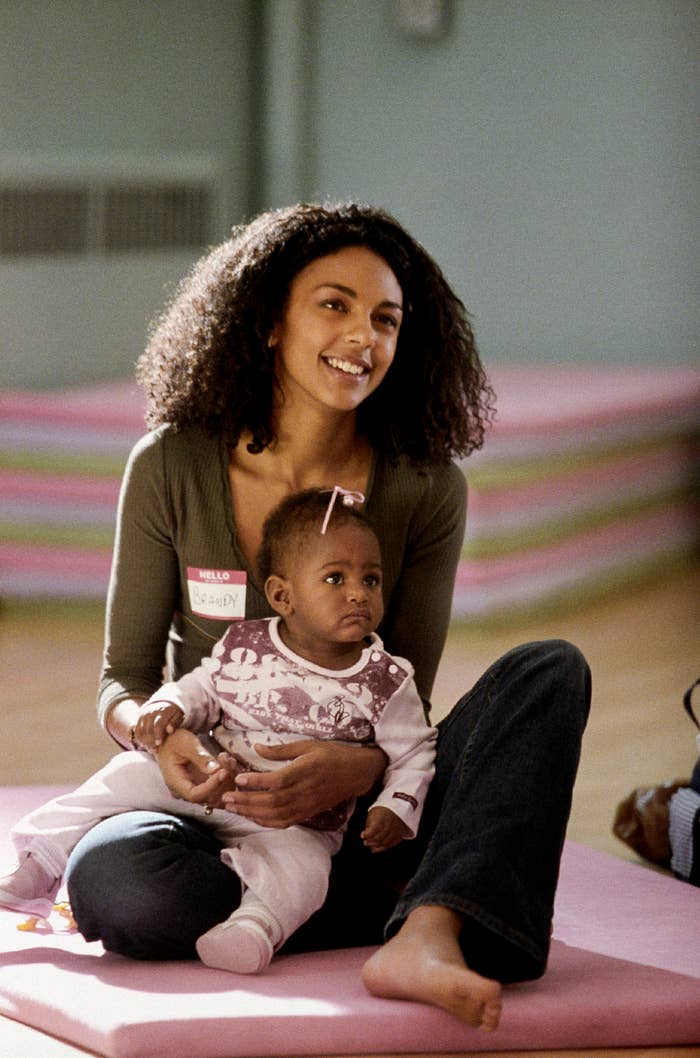 A woman, with a "Brandy" nametag, smiles while sitting on the floor holding a baby. She is dressed casually in jeans and a long-sleeve top