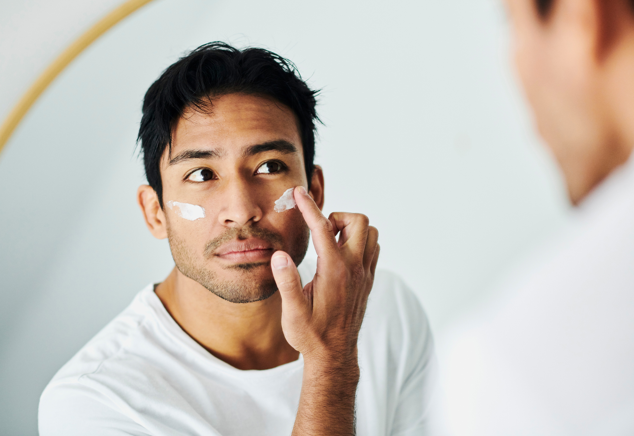 A man in a white shirt applies moisturizer to his face while looking into a mirror