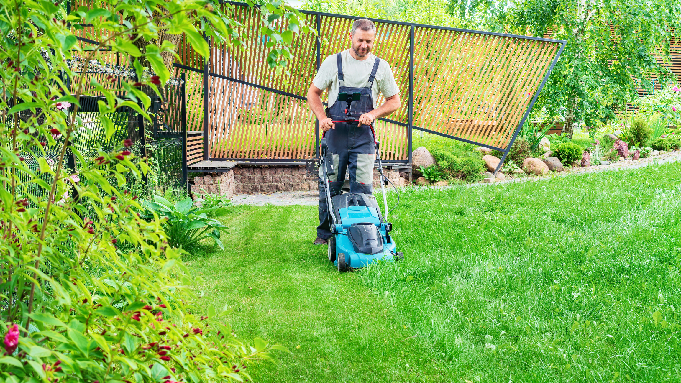 A person is mowing a lawn with an electric mower in a garden. The individual is wearing overalls and a white shirt