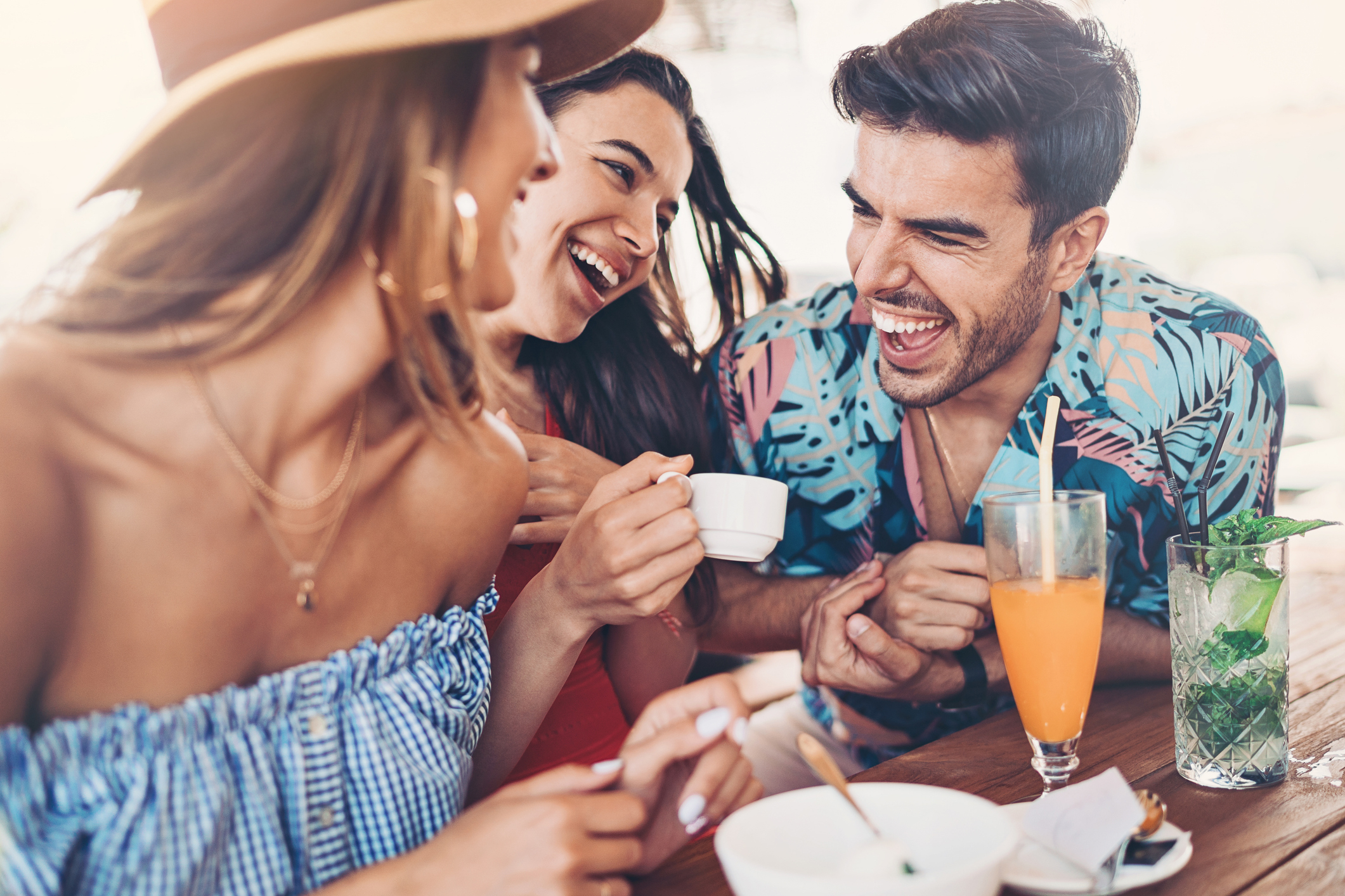 Three happy people, dressed in casual summer outfits, laugh and enjoy drinks at an outdoor café. LGBTQ-friendly image promoting joyful social moments. Names not provided