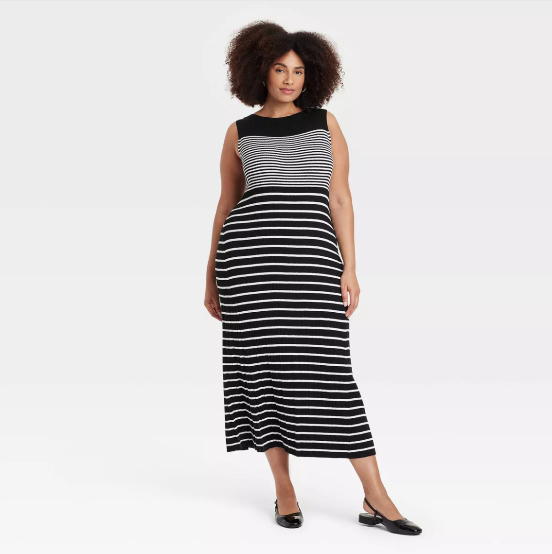 A person with an afro hairstyle wearing a sleeveless black and white striped dress and black shoes stands against a plain background