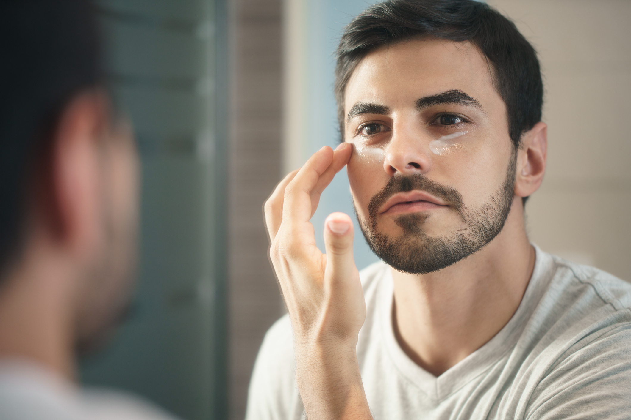 A man with short dark hair and a beard is applying skincare cream to his face while looking in a bathroom mirror. He is wearing a casual white t-shirt