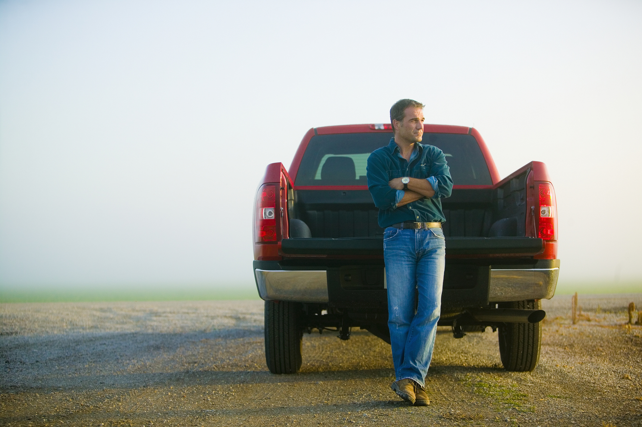 A man in casual attire leans against the back of a red pickup truck in a rural area on a foggy day