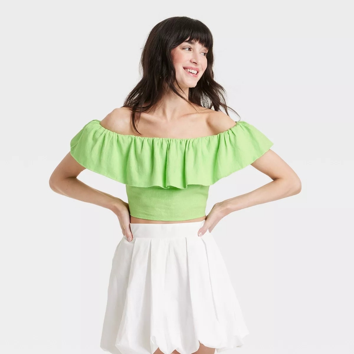 A model wears an off-shoulder ruffled top and a high-waisted skirt, smiling with arms on hips