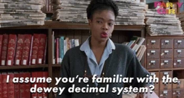 A scene from "Indie Film Girl" shows a person in a packed library speaking. Caption reads, "I assume you're familiar with the Dewey decimal system?"