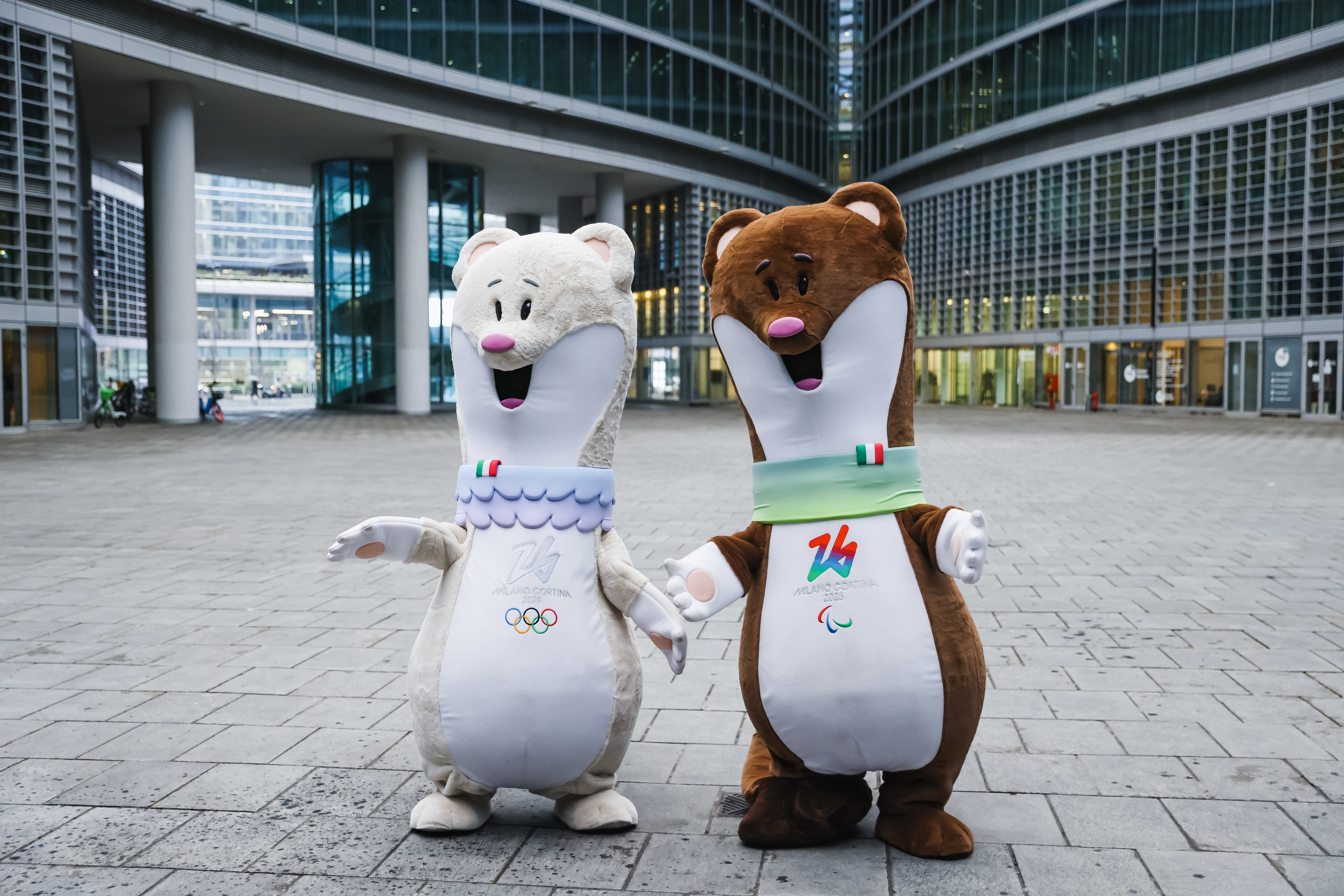 Two mascots, representing the Milan-Cortina 2026 Winter Olympics and Paralympics, stand in a plaza outside a modern building, waving and smiling