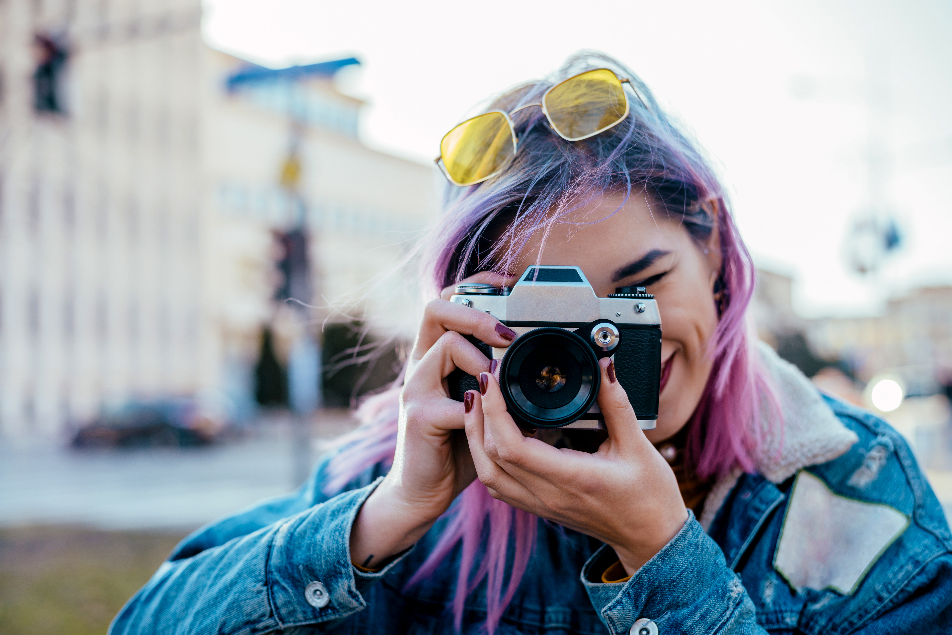 A person with dyed hair, wearing a denim jacket and yellow sunglasses, is holding and looking through a camera outdoors