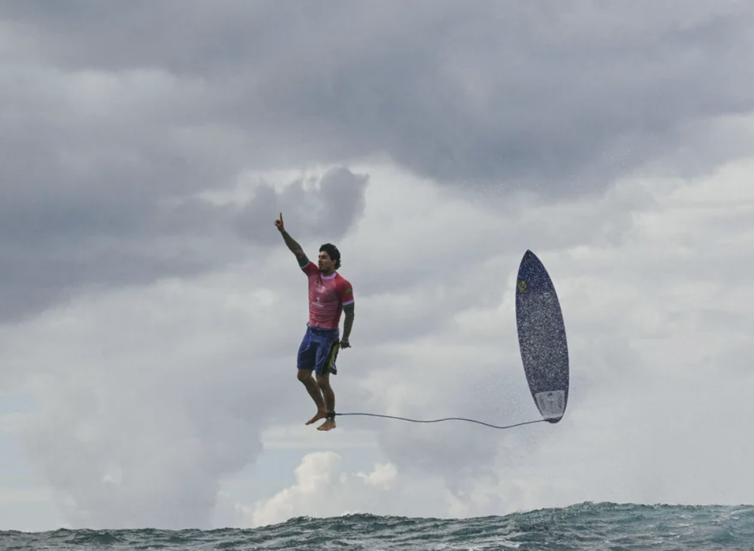 A surfer in mid-air above the ocean, pointing upwards with his right hand. A stormy sky is visible in the background. The surfboard is detached and to the right