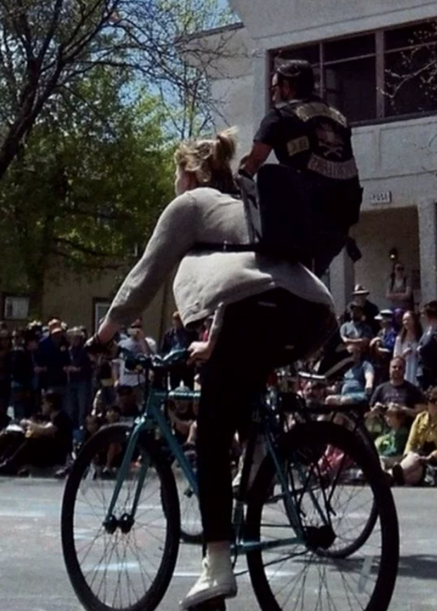 A man wearing a vest with patches rides on the back of a tandem bicycle being pedaled by a woman in a crowd. Trees and buildings are visible in the background