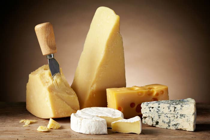 A variety of cheeses are displayed on a wooden surface, including a wedge of Swiss cheese, a wedge of blue cheese, a wheel of brie, and a partially cut block of Parmigiano-Reggiano with a cheese knife