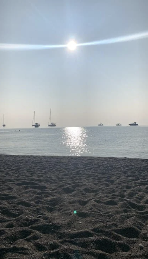 A calm beach scene at sunset, with the sun reflecting on the ocean. Five sailboats and yachts are anchored in the distance