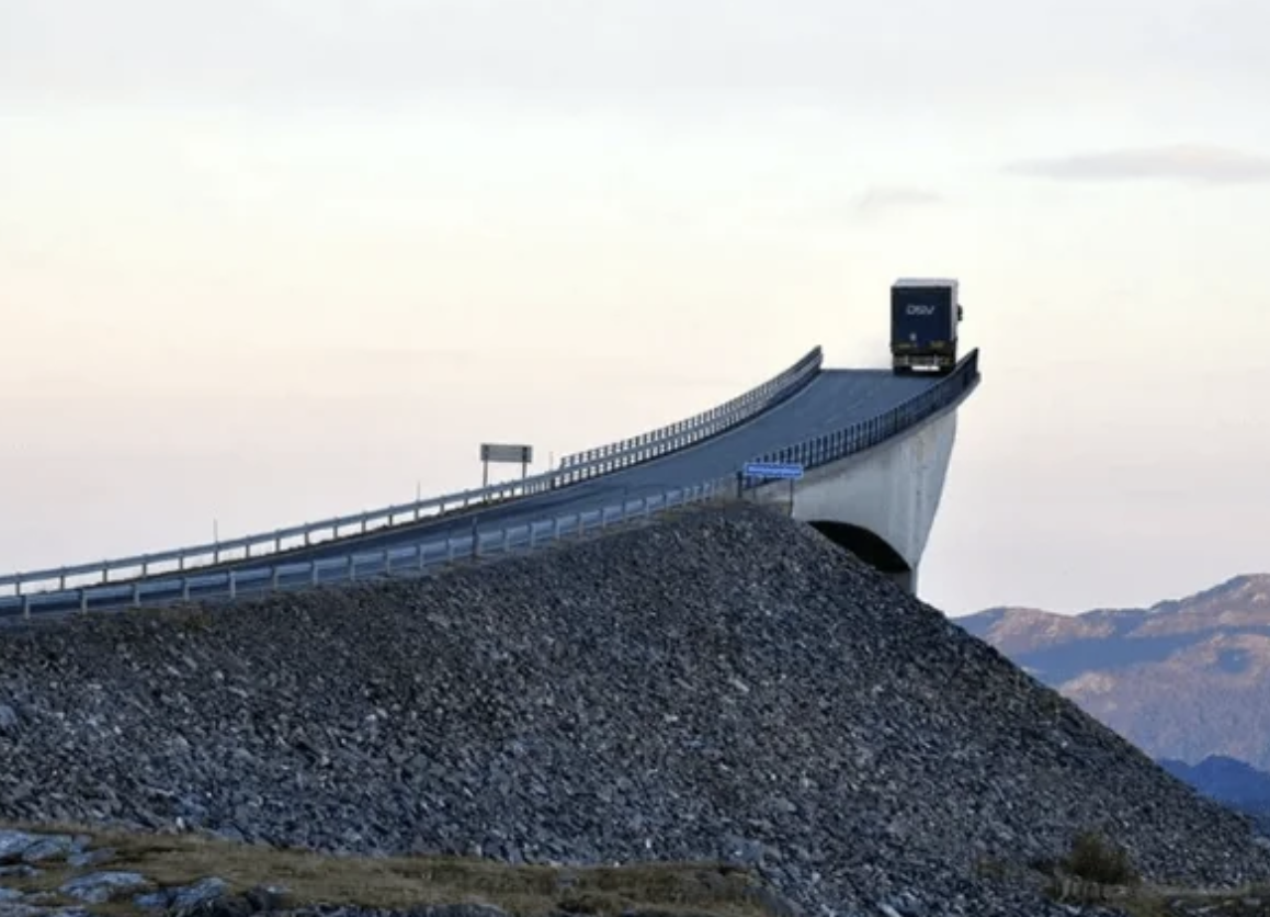 A large truck navigates a dramatic, steeply inclined bridge over rocky terrain with mountainous scenery in the background. No people are visible