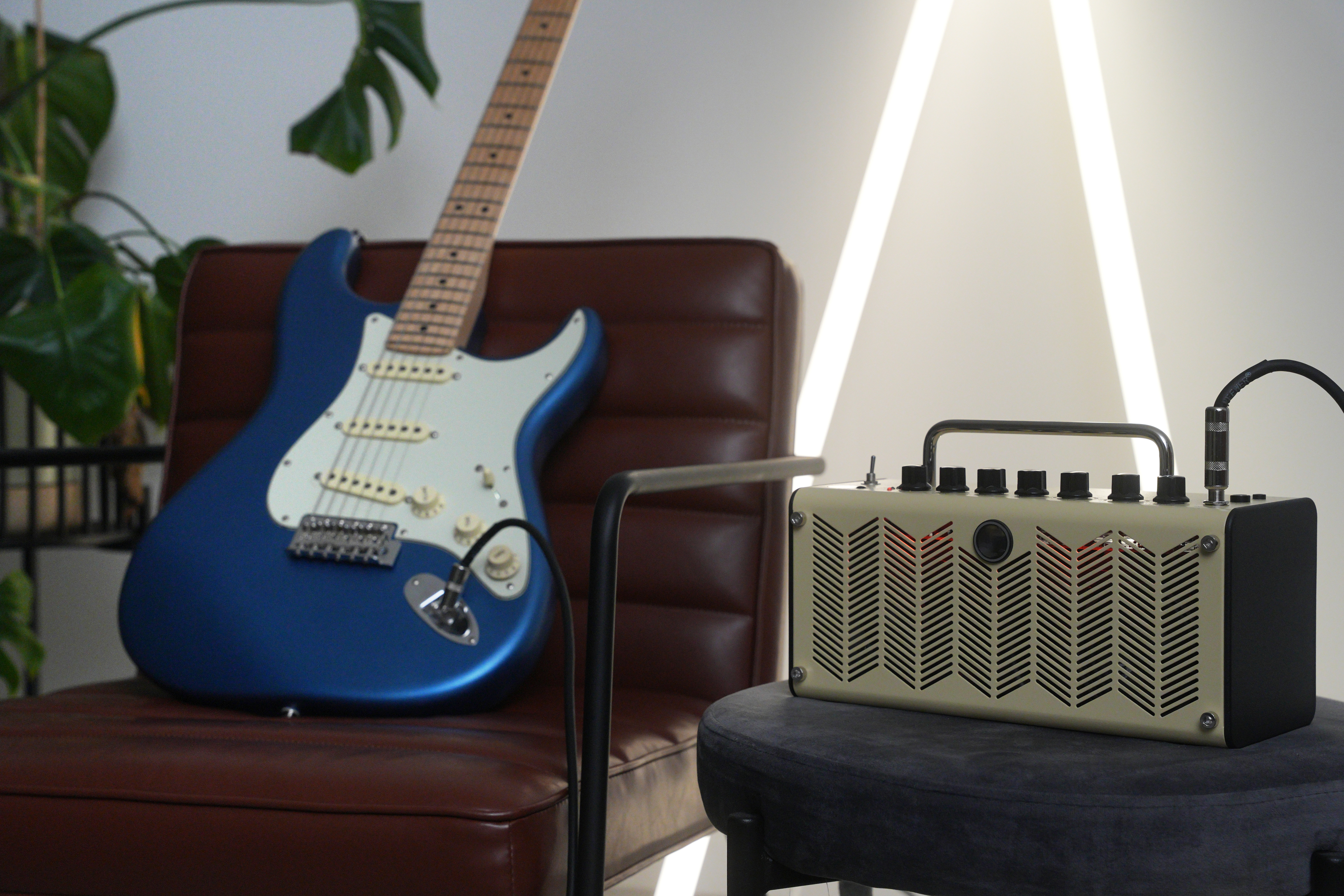 A blue electric guitar rests on a leather chair next to a small guitar amplifier placed on a stool. Both are set against a minimalist background with plants