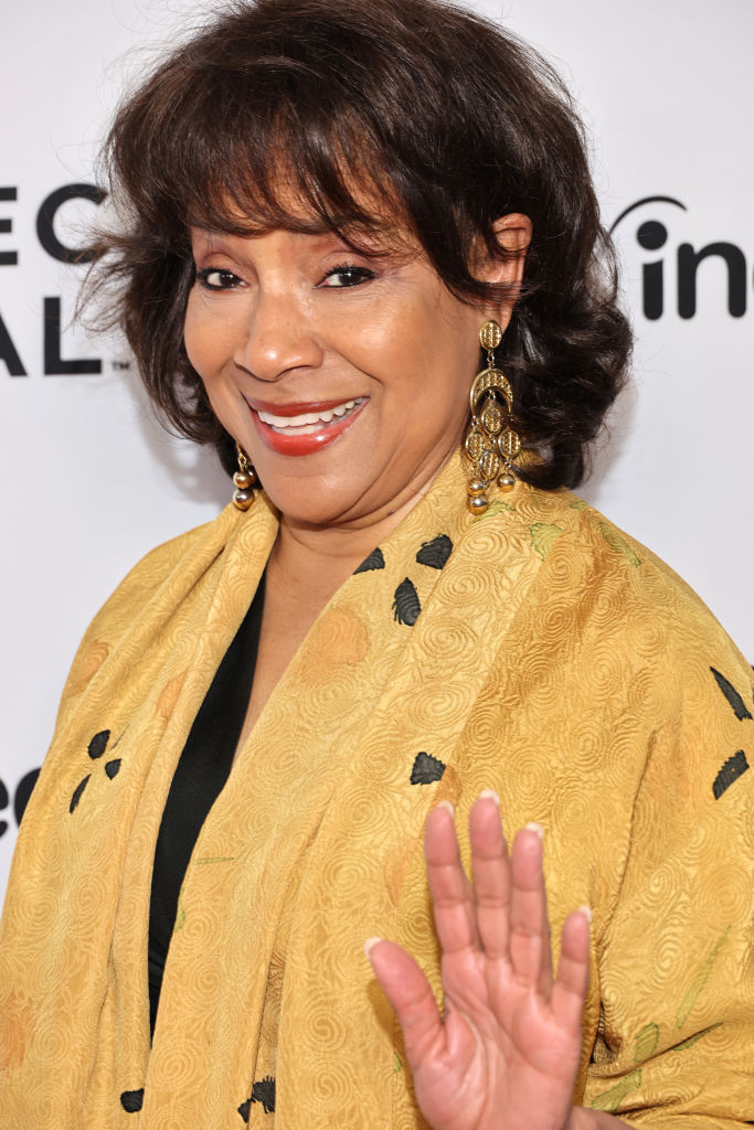 A woman smiles and waves at an event. She wears ornate gold earrings and a light shawl with a floral pattern