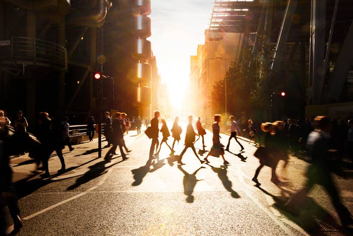 Silhouetted people walk across a city street at sunset, casting long shadows. Urban buildings and structures are visible in the background