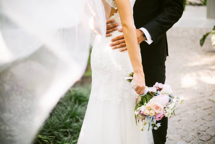 A bride and groom embrace at their wedding. The bride holds a bouquet of flowers while the groom's hand is placed on her back