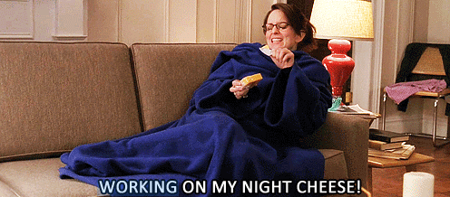 Tina Fey is sitting on a couch, wrapped in a blanket, eating cheese with the text &quot;WORKING ON MY NIGHT CHEESE!&quot; displayed