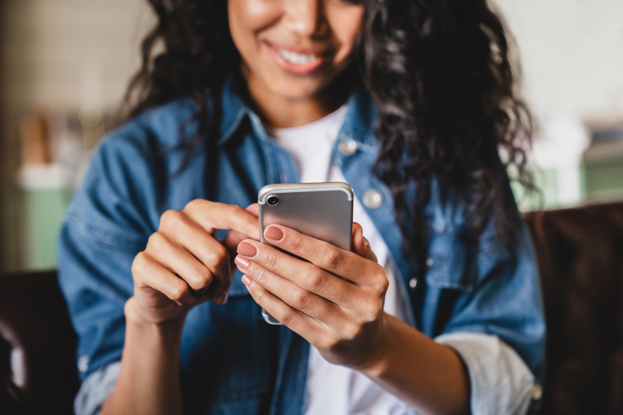 A person smiling and using a smartphone, wearing a casual denim jacket. The image is likely in an indoor setting. No other objects are visible