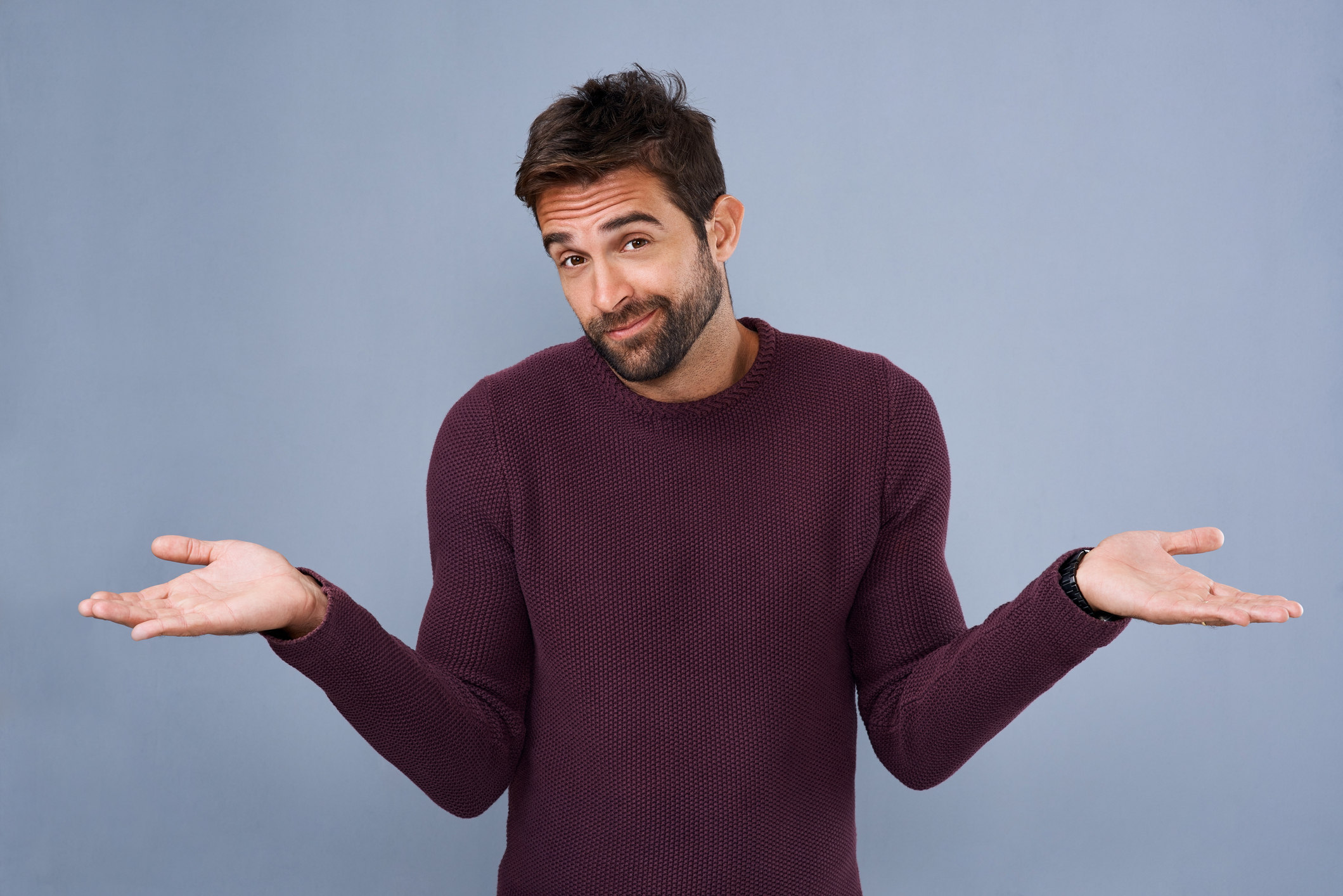 A man in a casual sweater stands against a neutral background, shrugging his shoulders with an uncertain expression