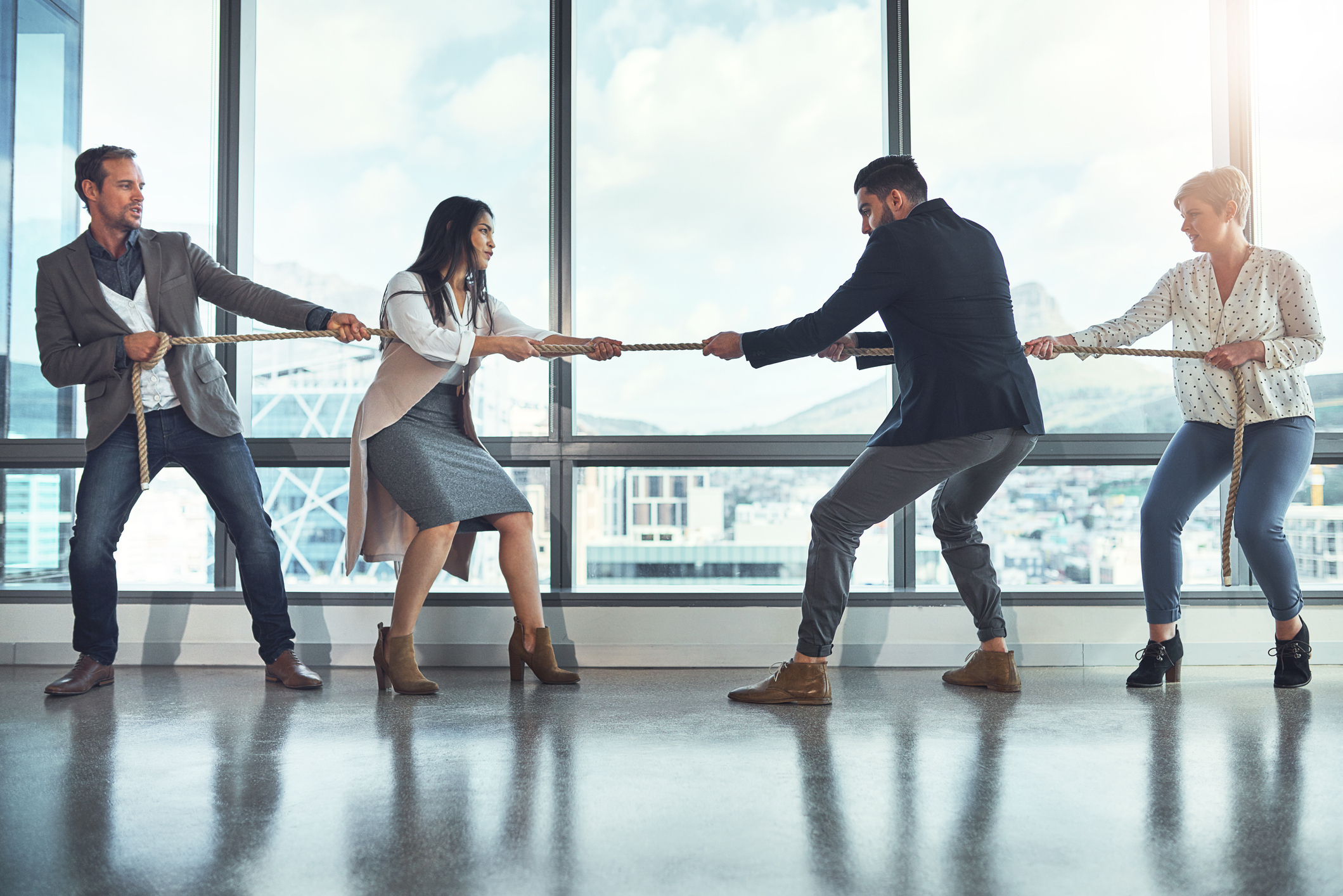 Four individuals, two men and two women, engage in an indoor tug-of-war near large windows overlooking a cityscape
