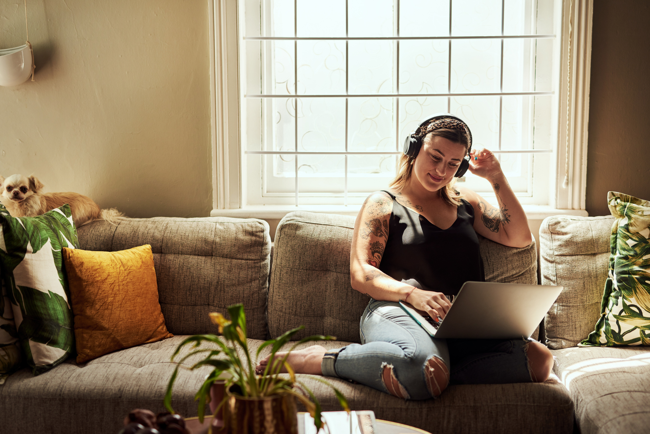 A woman with headphones sits on a couch using a laptop, with a small dog beside her and plants nearby