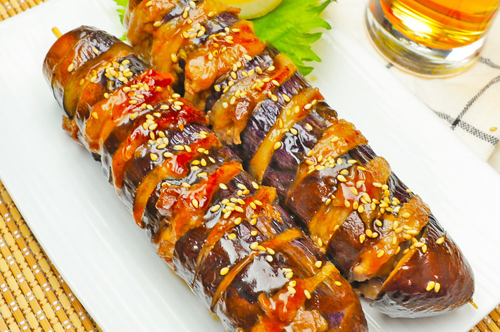 皿の上に並べられた2本のグリルされた茄子に、ゴマとソースがかかっています。右上にグラスが置かれています。