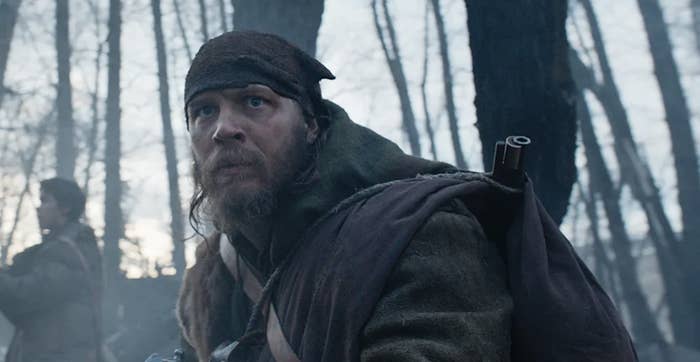 Tom Hardy dressed in rugged winter attire, wearing a headscarf, appears intense and focused in a forest setting. Another person is seen in the background
