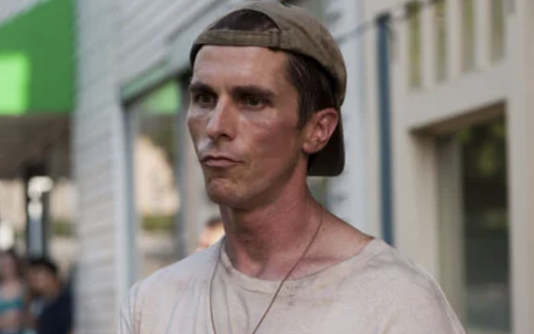 Christian Bale, wearing a backward cap and a worn-out t-shirt, standing outside with a neutral expression