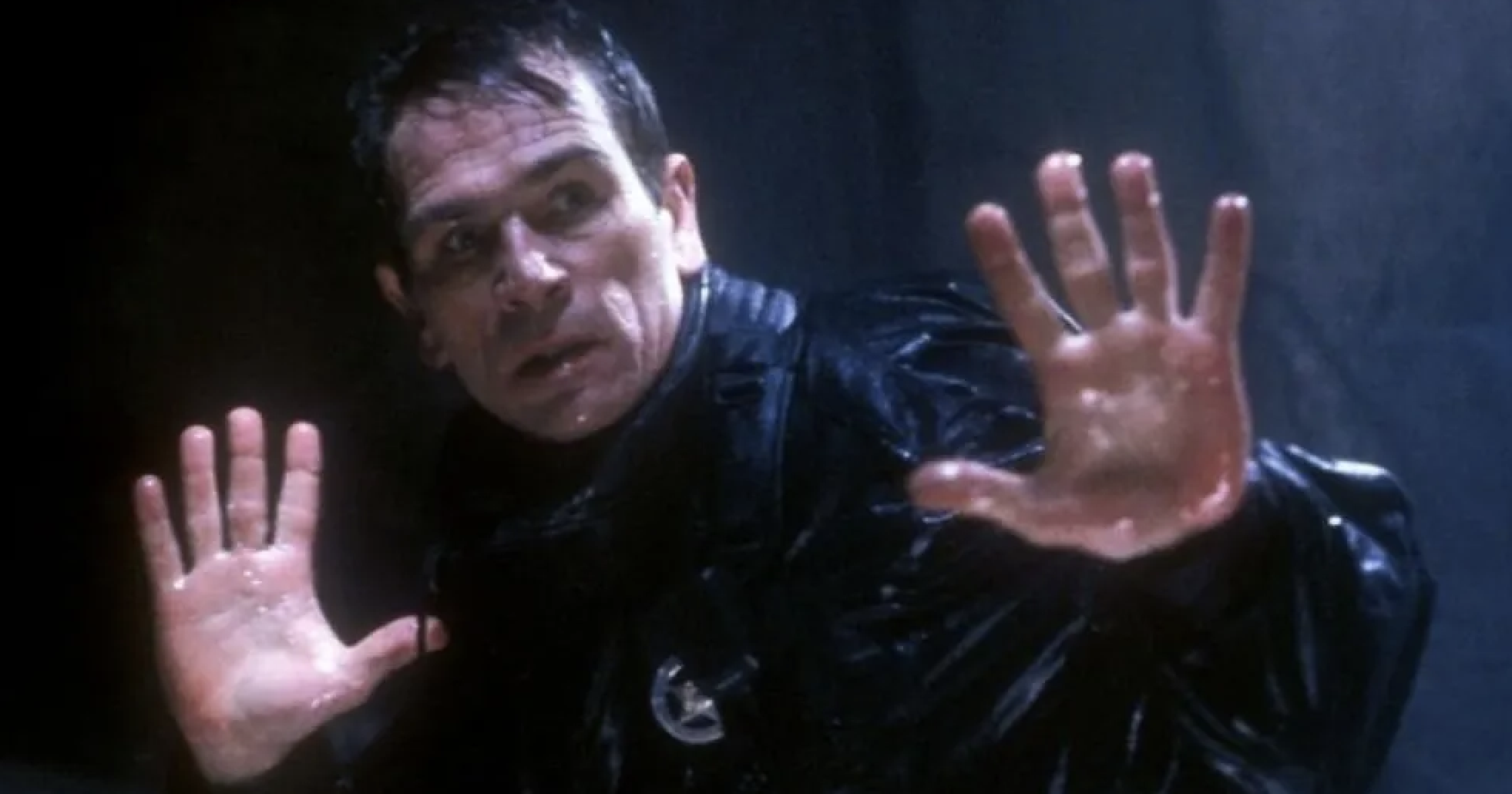 Man in wet black clothing with raised hands, appearing nervous or alert in a dimly lit scene
