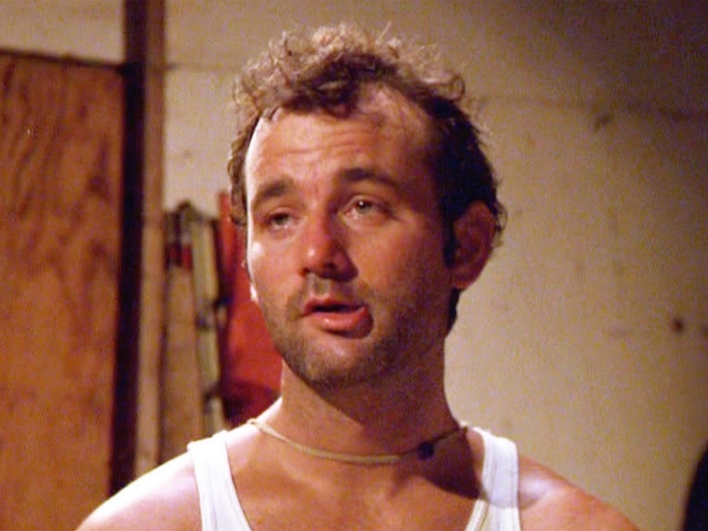 Bill Murray appears disheveled in a white tank top and necklace, looking tired in an indoor setting