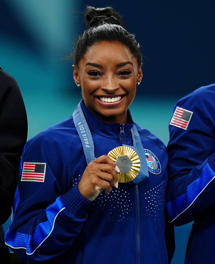 Simone Biles smiles holding a gold medal wearing an athletic team jacket with USA flag patches