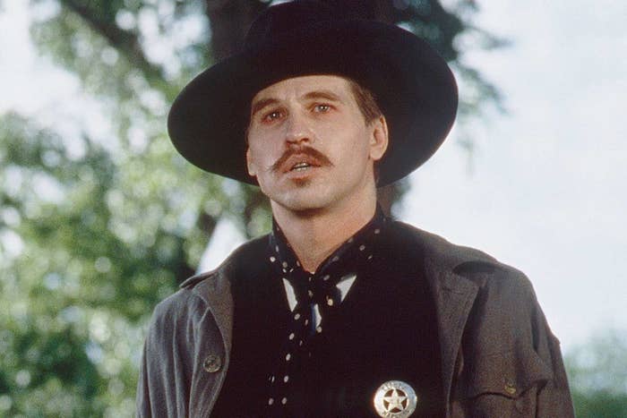 Val Kilmer as Doc Holliday in western attire, including a wide-brimmed hat and a sheriff's badge
