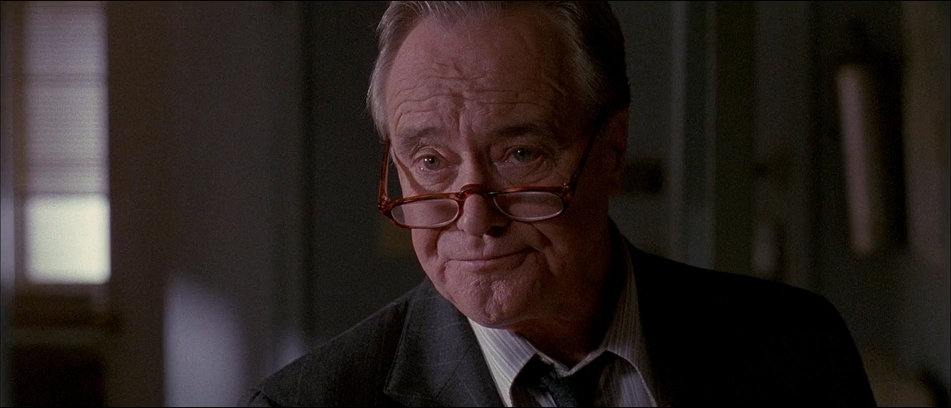 An older gentleman in a suit and tie, wearing glasses, is seen with a slight smile and raised eyebrows suggesting skepticism or amusement. Names not known