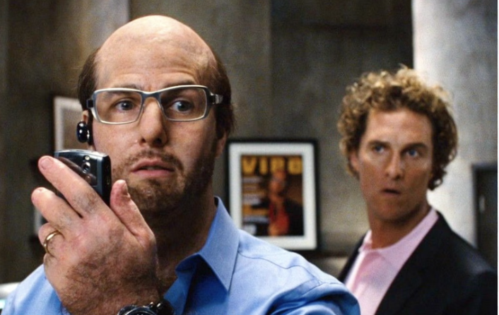Tom Cruise, wearing a bald cap with glasses, holds a device to his mouth; Matthew McConaughey, in a suit, looks surprised in the background