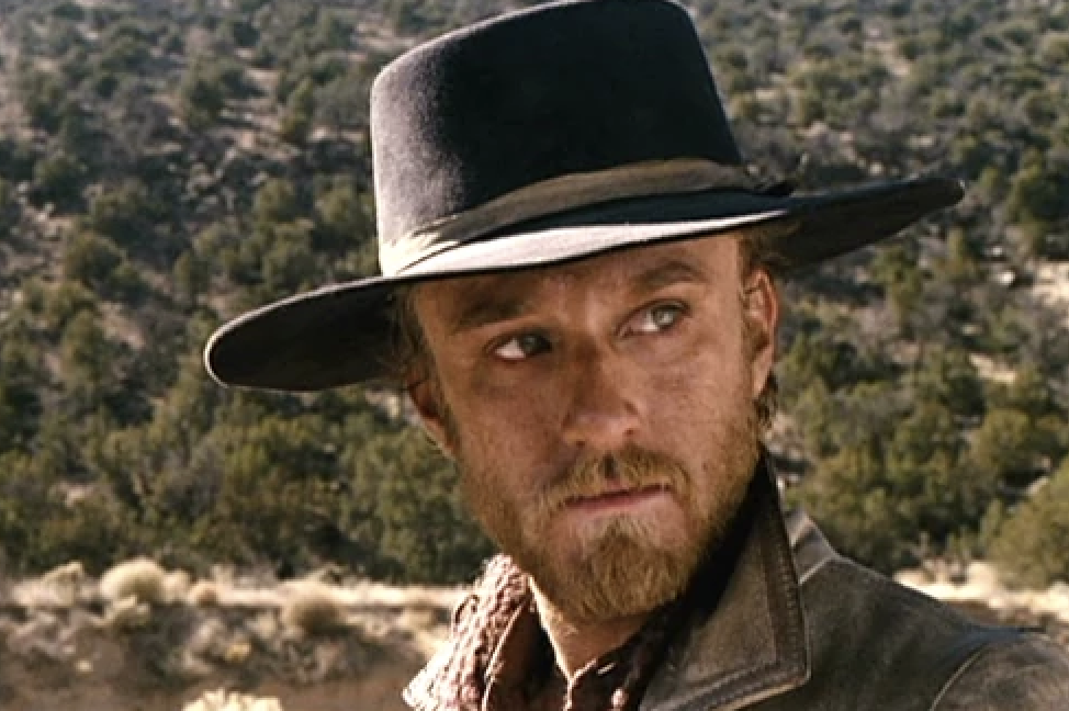 A person wearing a wide-brimmed hat and a leather jacket stands outdoors in a rugged, natural setting