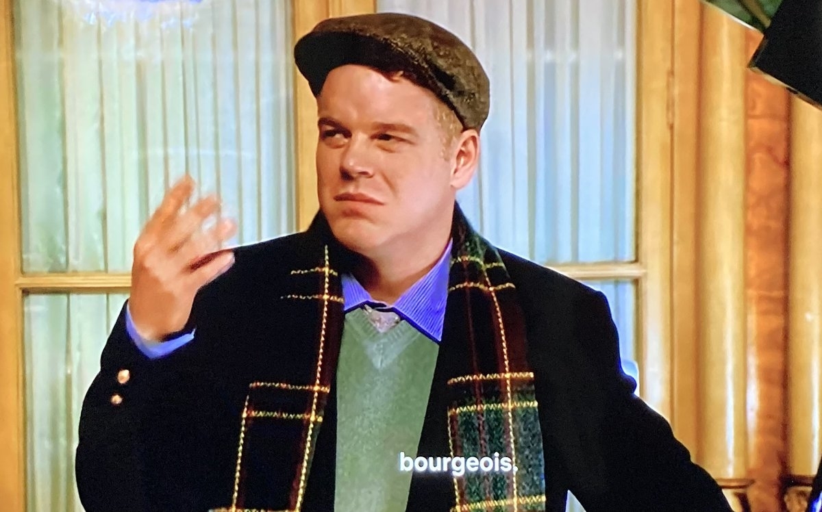 A man wearing a flat cap, a blazer, a sweater, a collared shirt, and a scarf is standing indoors. The subtitle reads "bourgeois."