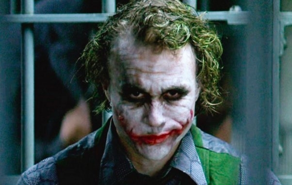 Heath Ledger as the Joker, with green hair and white face makeup, wearing a gray shirt and a green vest, smirking behind bars in a dim setting
