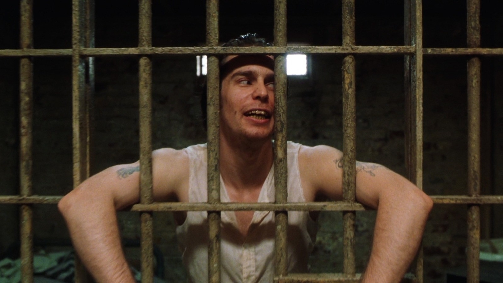 Man with tattoos on arms and wearing a sleeveless shirt looks through prison bars with a smirk on his face