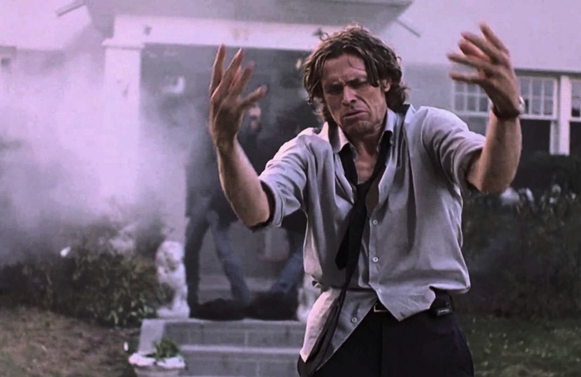 Willem Dafoe stands outstretched, looking distressed in front of a building with smoke and another person in the background