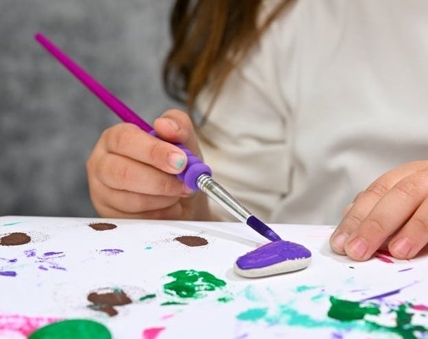 A young child is painting a small stone with a brush, surrounded by various painted stones and craft materials on the table
