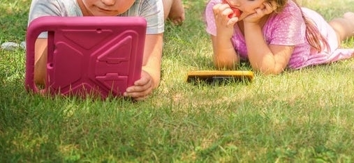 Two children lying on grass, focused on tablet devices. The boy holds a tablet with a pink case, and the girl has her device resting in front of her