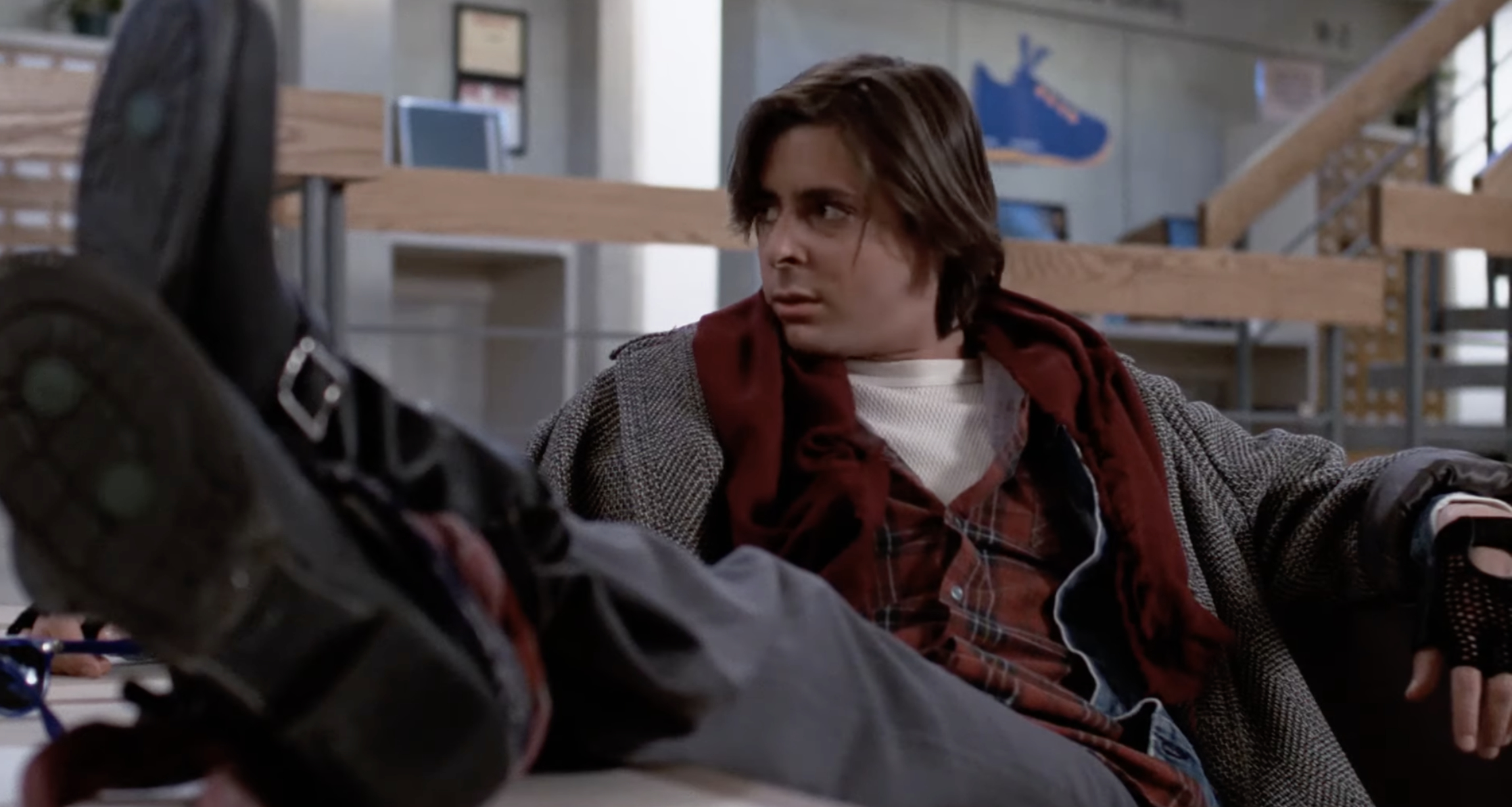 A teenager wearing mismatched layers of clothing and boots lounges on a table in what appears to be a school setting, looking off to the side