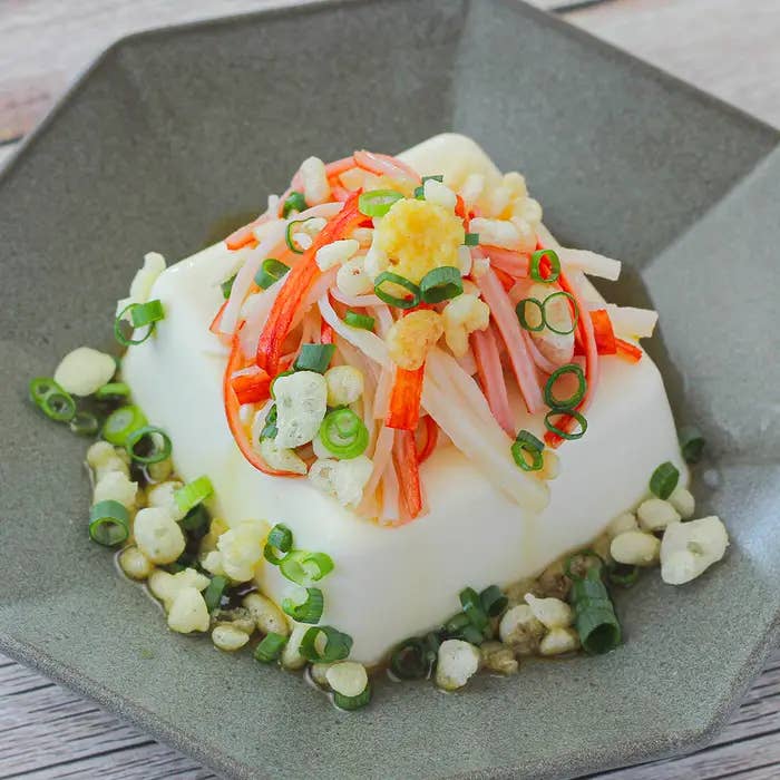 皿に盛られた豆腐の上に細かく刻まれたカニかまぼこ、ネギ、天かすがトッピングされている料理。
