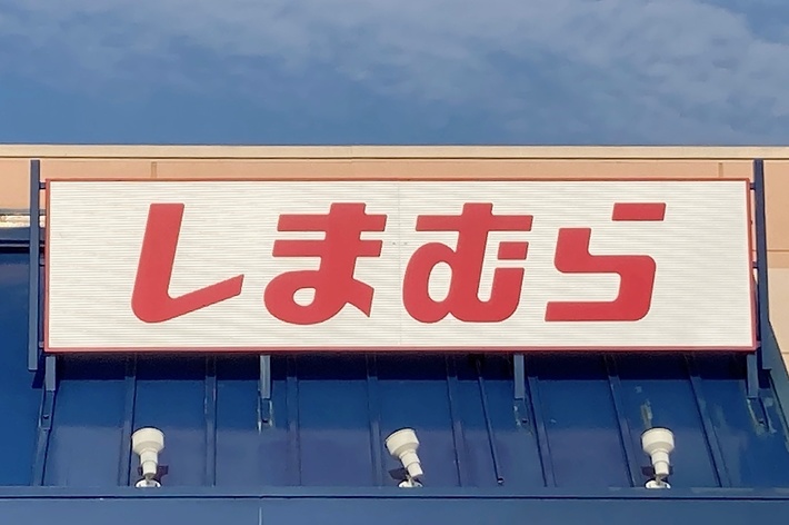 Storefront with a sign in Japanese, which reads &ldquo;しまむら&rdquo; (Shimamura). The roof is blue, and a disabled parking sign is visible below