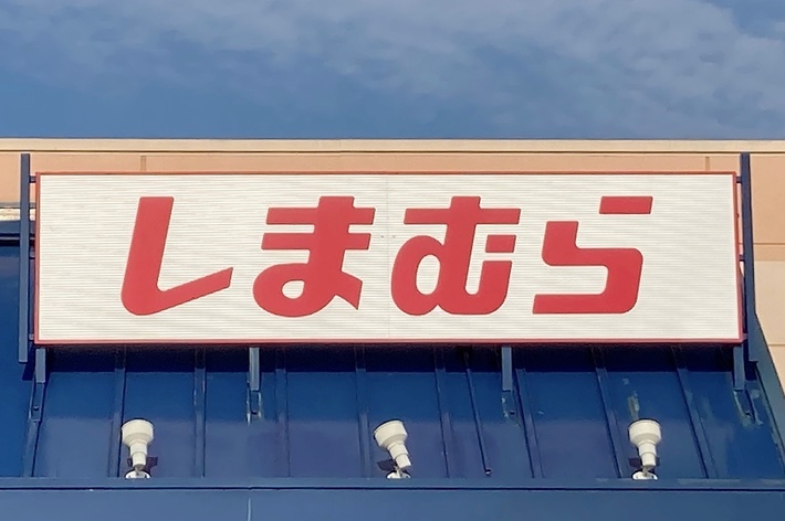 Sign with red Japanese characters on a storefront roof reads &ldquo;しまむら&rdquo;. Posts with four white lamps are visible below the sign