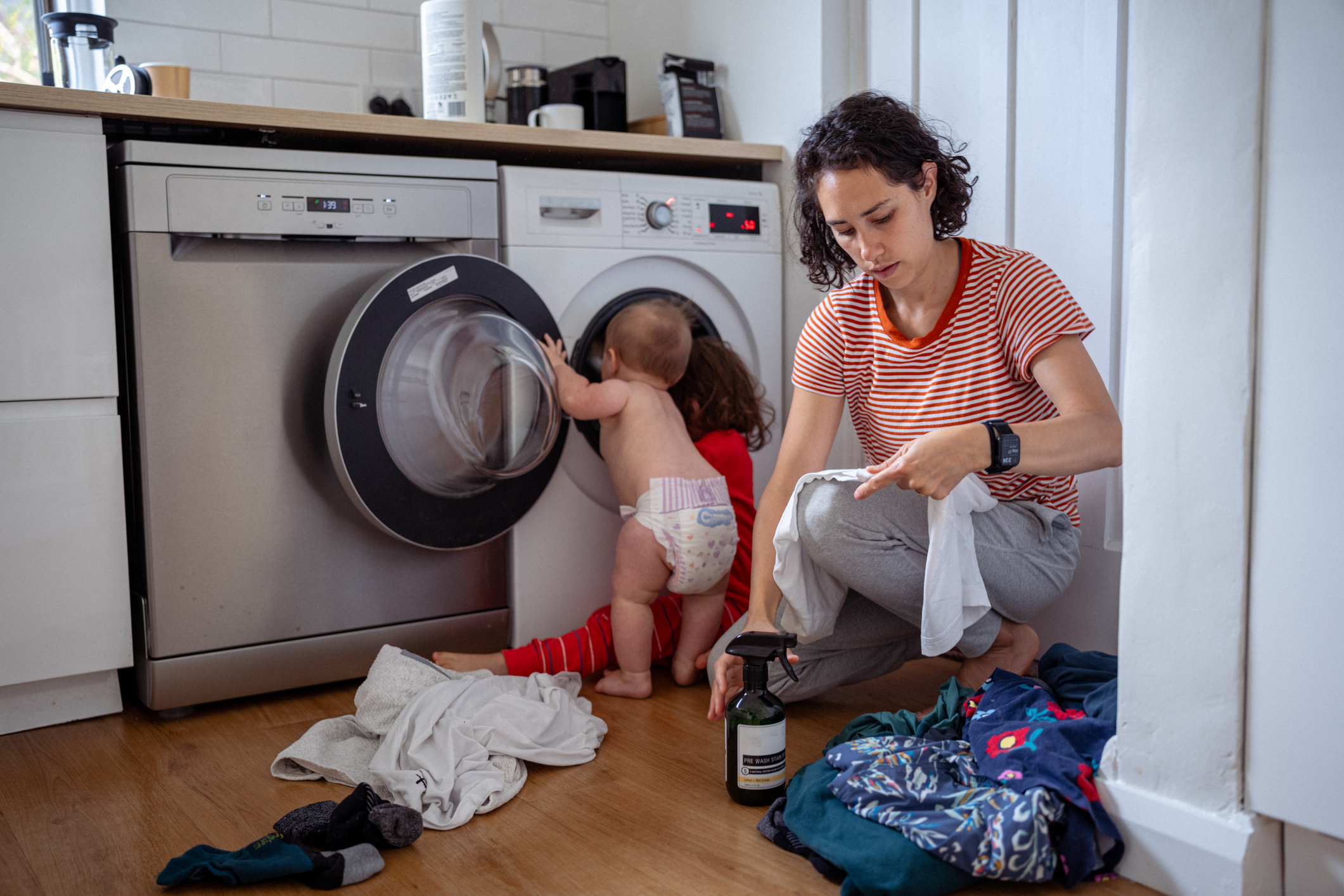 A woman in a striped shirt sits beside a baby in a diaper who is exploring the inside of a washing machine. Laundry is scattered around them