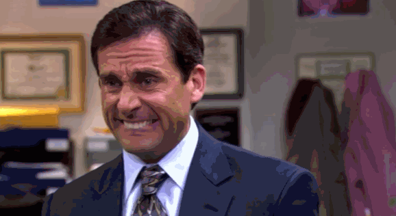 Steve Carell in an Office setting, making a cringing facial expression while wearing a suit and tie. Wall frames and coats are blurred in the background