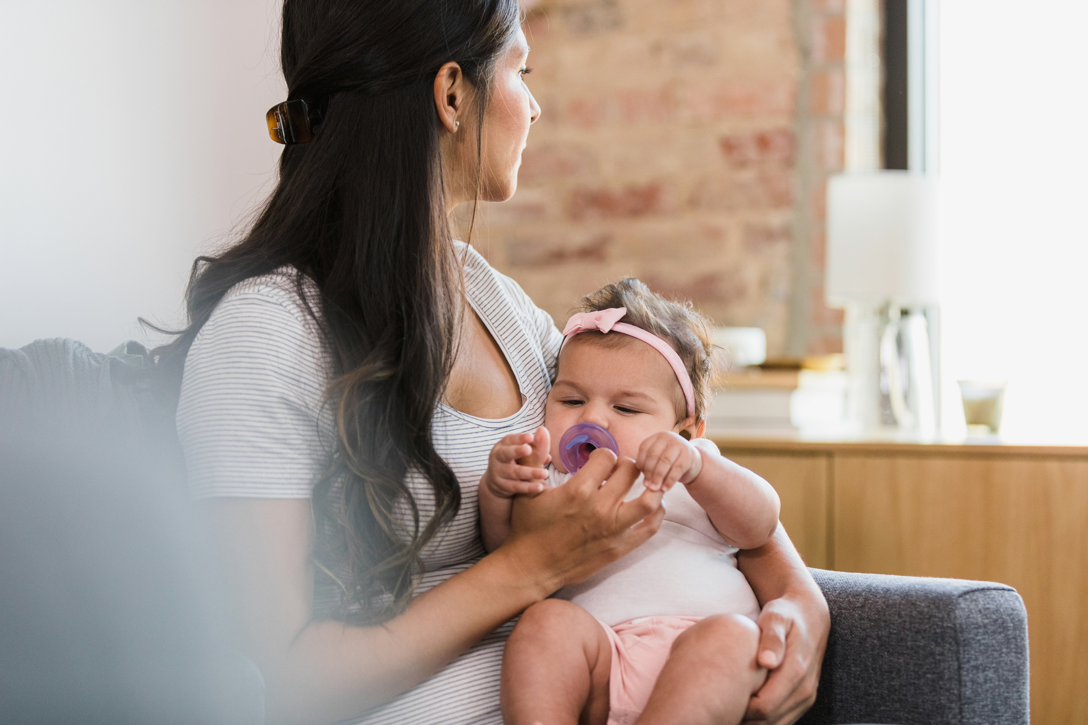 A woman is sitting and holding a baby with a pacifier. The baby is wearing a headband and onesie. The woman has long hair and is looking away from the camera